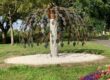 Metal tree sculpture with heart-shaped leaves stands on a white gravel circle in a park.