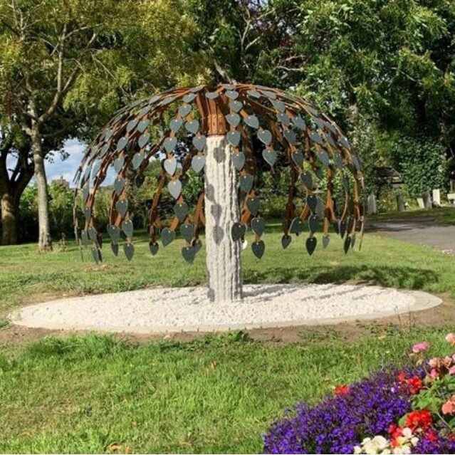 memorial_tree Metal tree sculpture with heart-shaped leaves stands on a white gravel circle in a park.