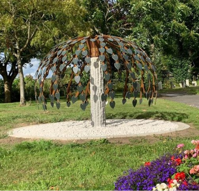 memorial_tree Metal tree sculpture with heart-shaped leaves stands on a white gravel circle in a park.