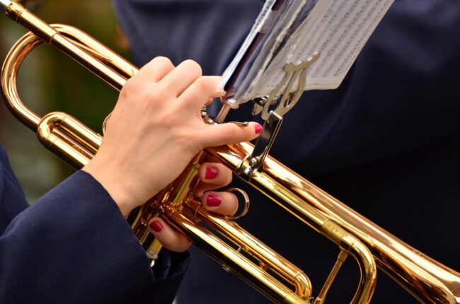 trumpet-3303724_1280 Close-up of hands with red nails playing a golden trumpet, sheet music visible above.