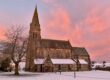 A stone church with a tall spire, snowy ground, and a pink sunset sky in the background.