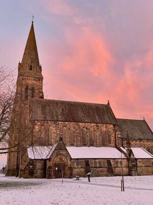 IMG_1960 A stone church with a tall spire, snowy ground, and a pink sunset sky in the background.