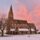 A stone church with a tall spire, snowy ground, and a pink sunset sky in the background.