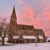 A stone church with a tall spire, snowy ground, and a pink sunset sky in the background.