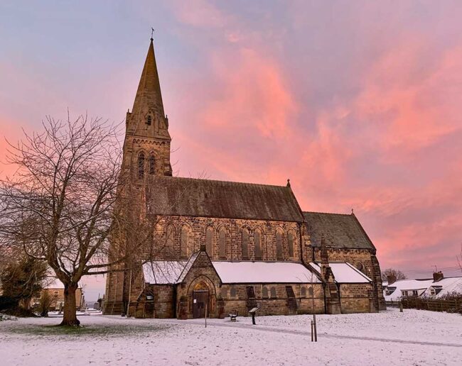 IMG_1960 A stone church with a tall spire, snowy ground, and a pink sunset sky in the background.