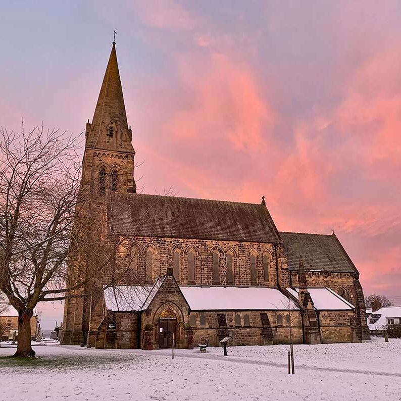IMG_1960 A stone church with a tall spire, snowy ground, and a pink sunset sky in the background.