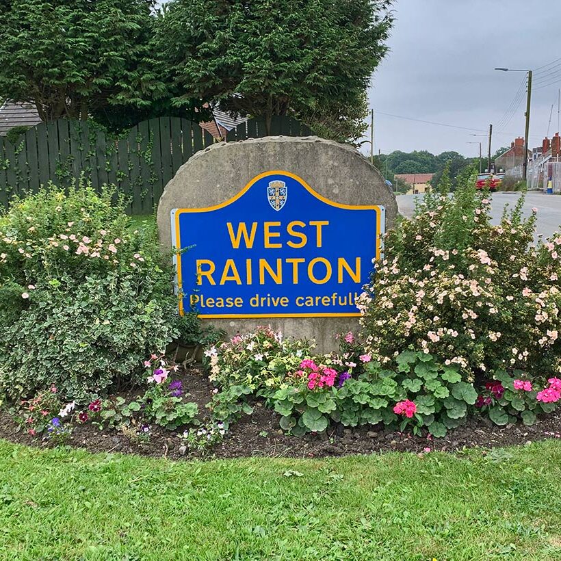 A blue West Rainton sign surrounded by flowers and bushes beside a road.