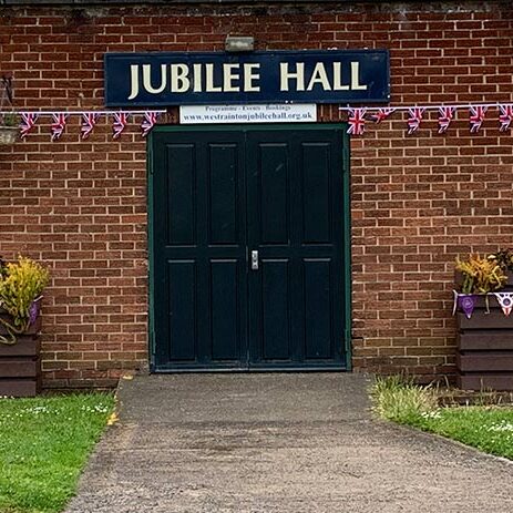 Double doors of Jubilee Hall with bunting, flower planters, and a defibrillator on a brick wall.